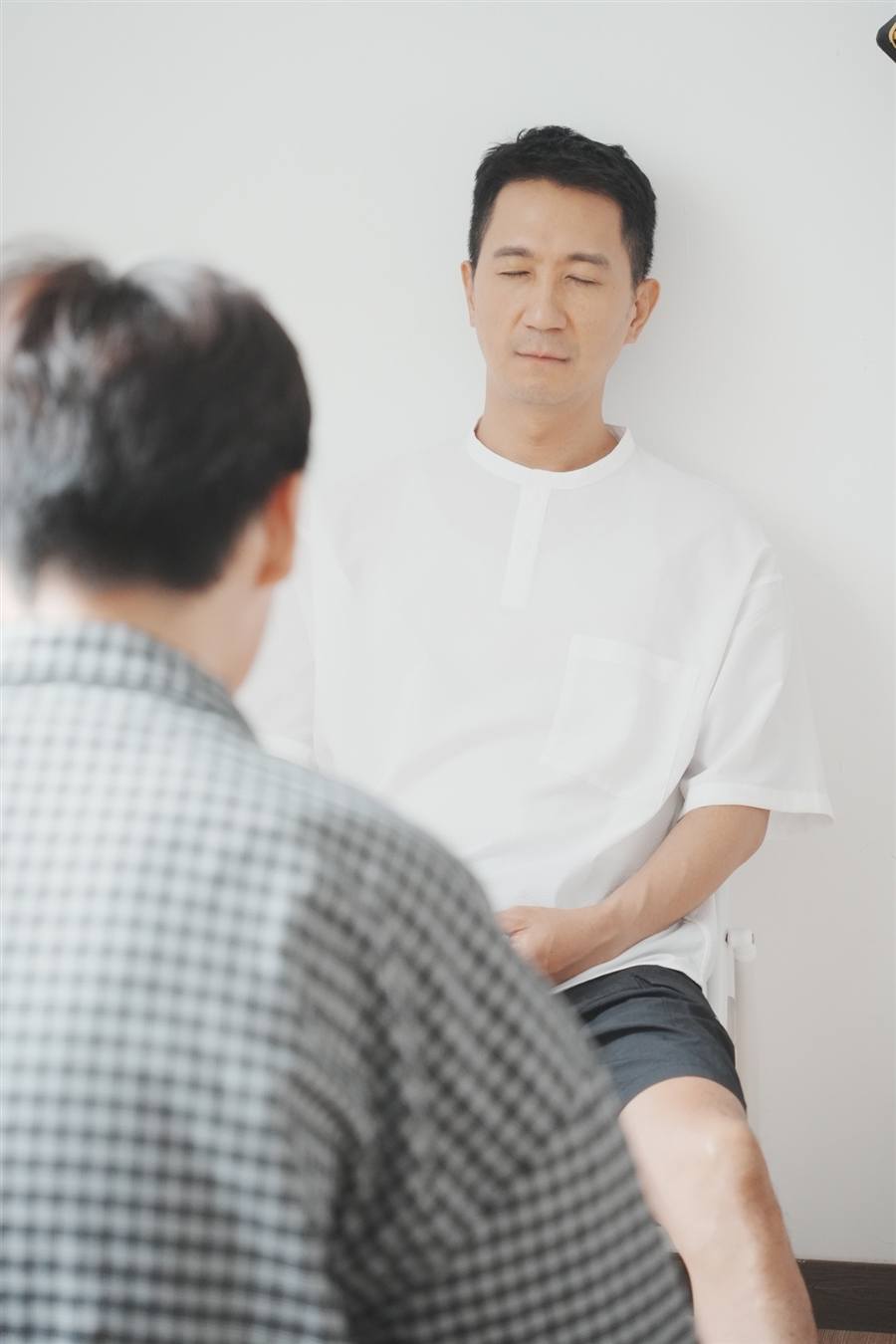 一個人靠著白牆坐著、閉著眼睛,另一個人在前景陪著